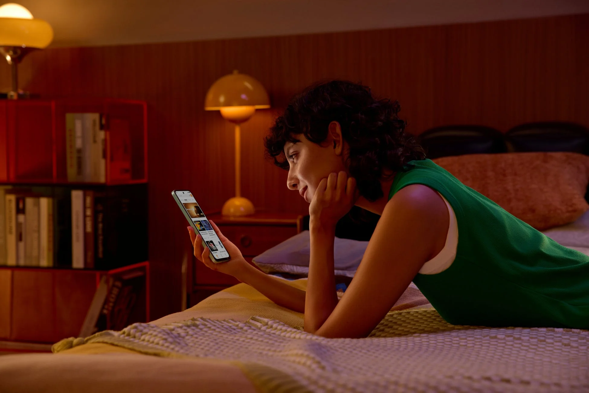 温かい照明の部屋で、ベッドに横たわる女性がスマートフォンを操作している。リラックスした様子で、デジタルコンテンツを楽しんでいるようだ。